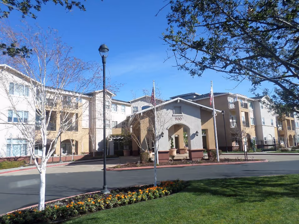 Exterior view of a multi-story senior living facility building with a driveway, landscaped flower beds, trees, and two flagpoles in front under a clear blue sky.