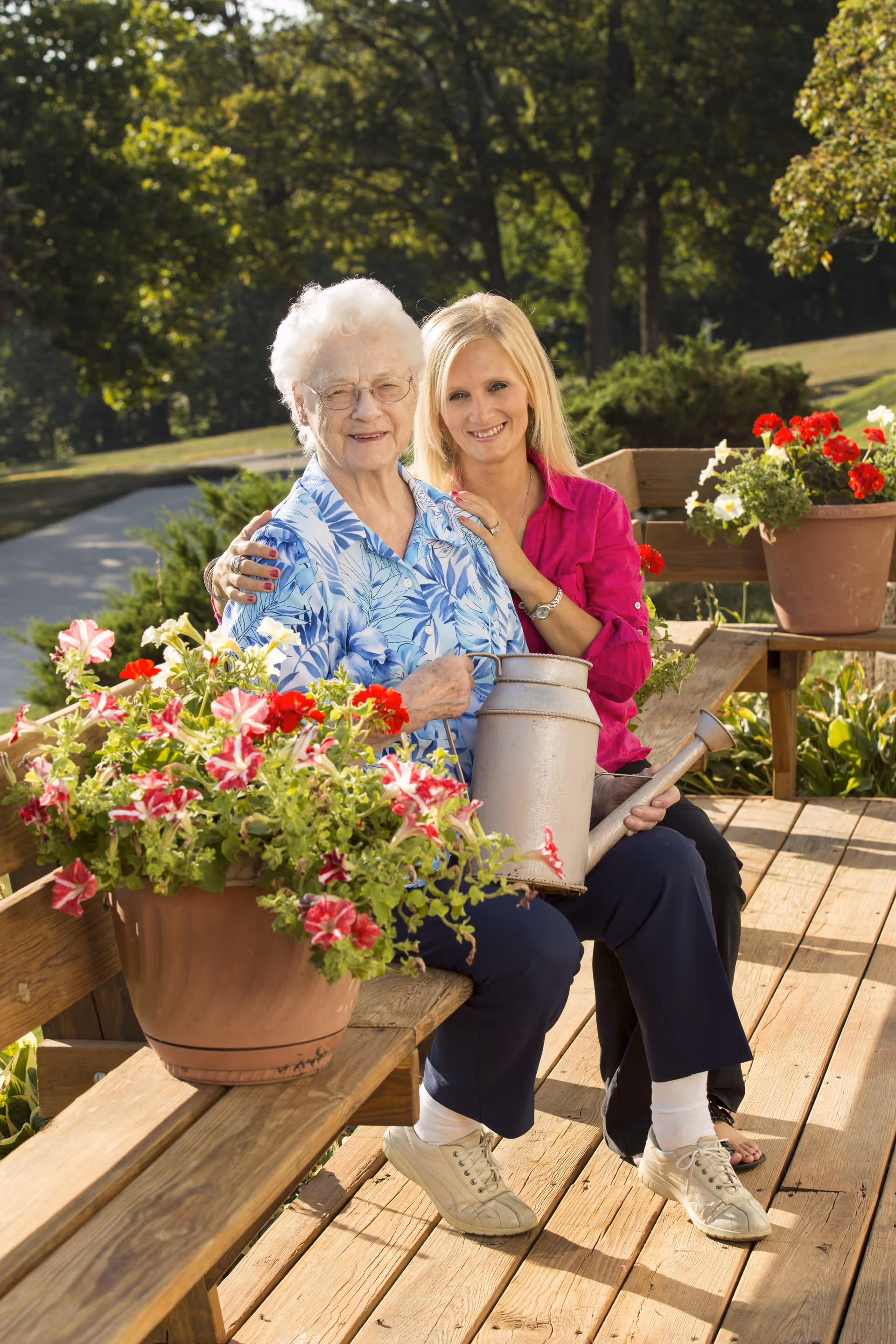 An elderly woman and a younger woman sitting together on a wooden bench outdoors surrounded by potted flowers. The elderly woman is holding a watering can, and both are smiling at the camera with greenery and trees in the background.
