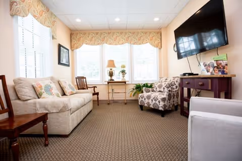 Cozy senior living lounge with sofas, chairs, TV, windows, and patterned carpeting