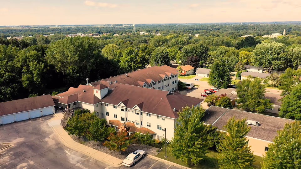 Aerial view of Pleasant View Estates, a multi-story senior living facility surrounded by trees and greenery, with a parking lot and several cars visible in front of the building.