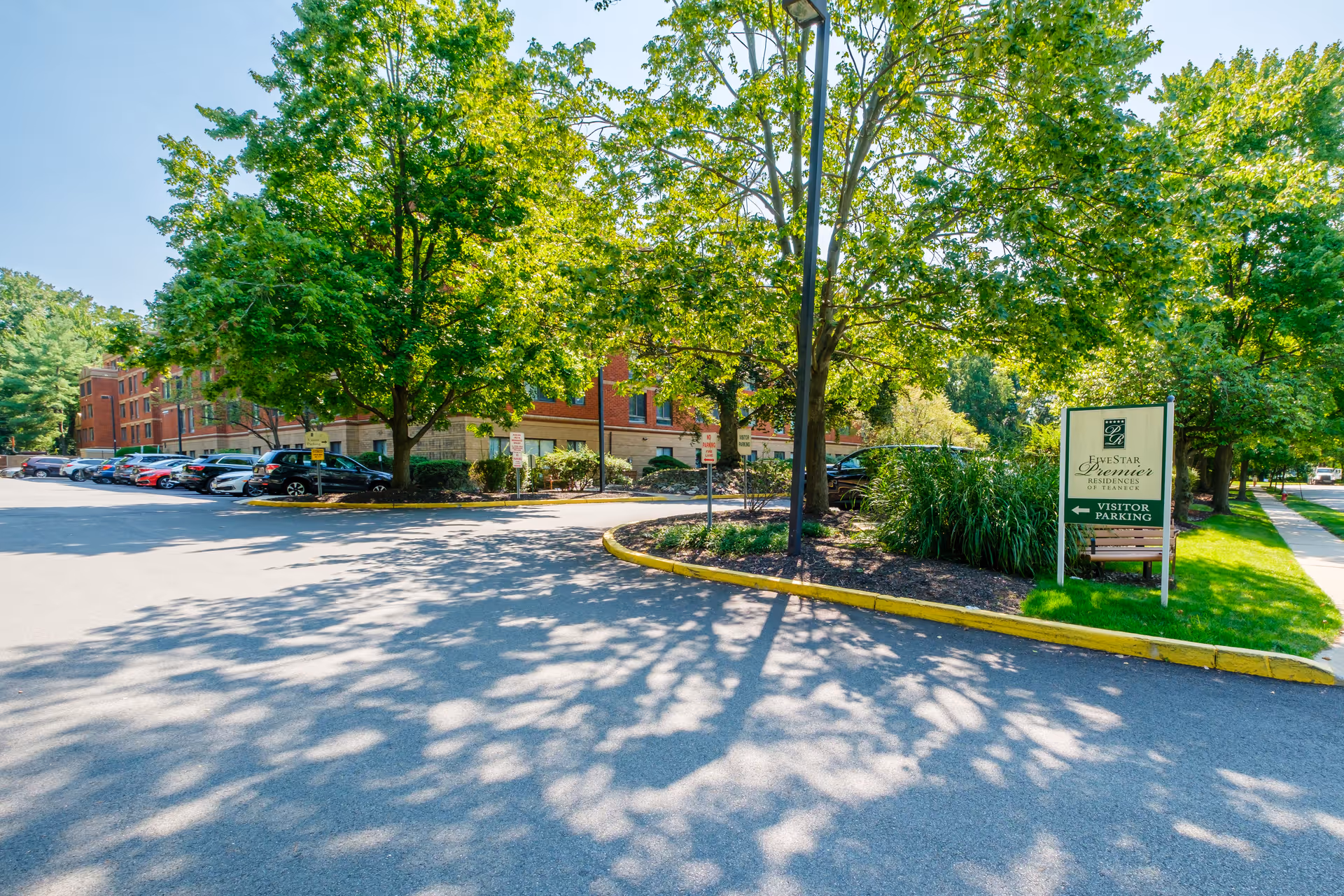 Exterior view of a brick senior living building with a tree-lined driveway, parked cars, and a 'Visitor Parking' sign for Five Star Premier Residences.