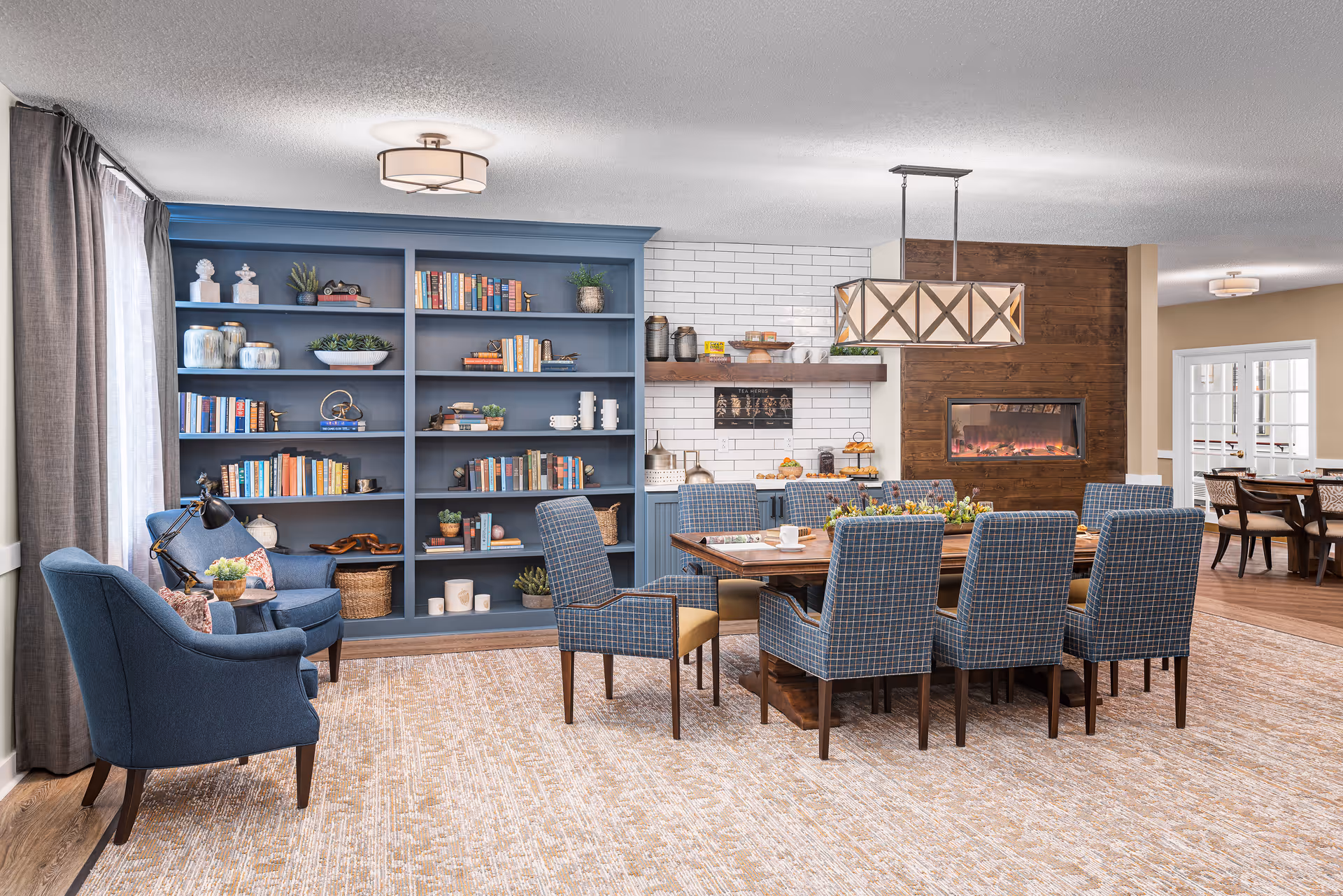 A cozy and well-lit common area featuring a large wooden dining table surrounded by eight blue upholstered chairs with a checkered pattern. To the left, there are two blue armchairs with a small side table and a lamp between them. Behind the armchairs is a large blue bookshelf filled with books, decorative items, and plants. The back wall has white subway tiles, a wooden shelf with various items, and a modern electric fireplace set in a wooden accent wall. In the background, another dining area with a table and chairs is visible through an open doorway.