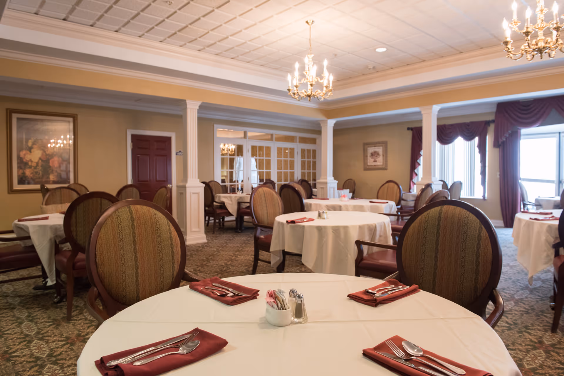 Formal dining room with round tables covered in white tablecloths, set with red napkins and chandeliers overhead.