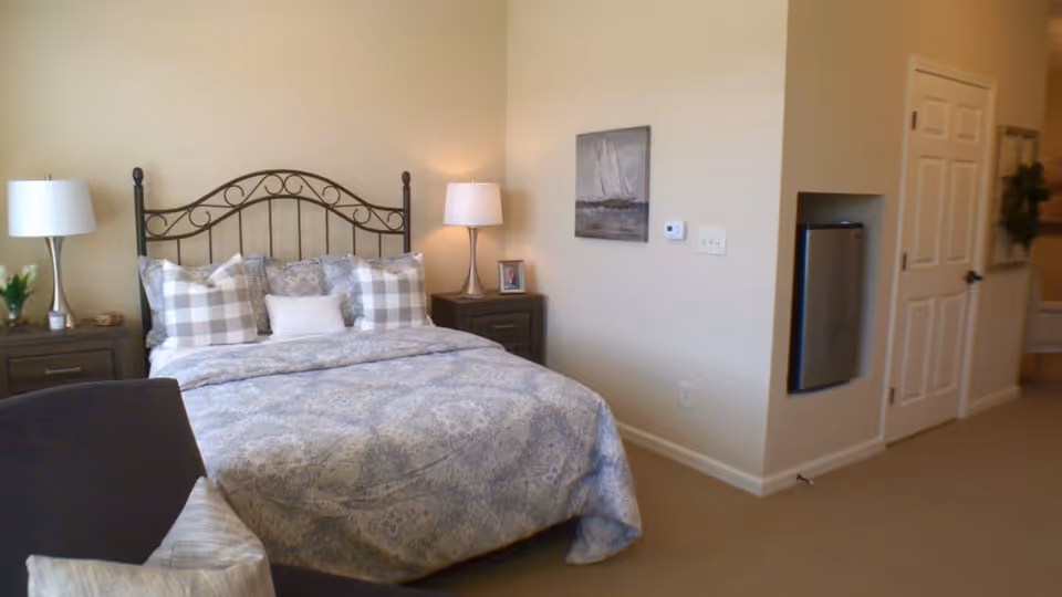 A cozy bedroom with a metal headboard bed dressed in patterned bedding and multiple pillows. There are two matching nightstands on either side of the bed, each with a lamp. A small framed photo and a decorative plant are on the nightstands. A painting of a sailboat hangs on the wall above a thermostat. A mini refrigerator is built into the wall near a closed white door. The room has beige walls and carpeted flooring.