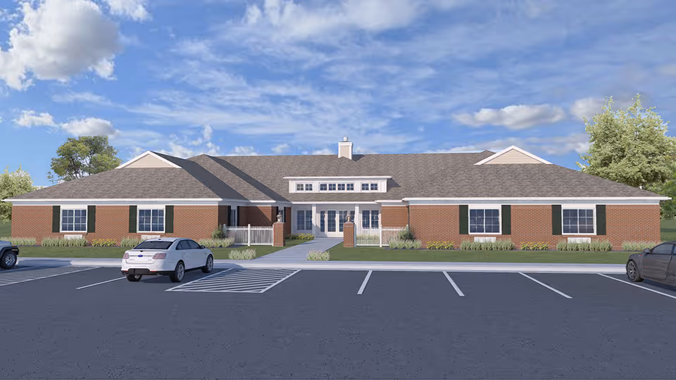 Single-story brick senior living building with a central entrance, landscaped beds and a parking lot under a blue sky.