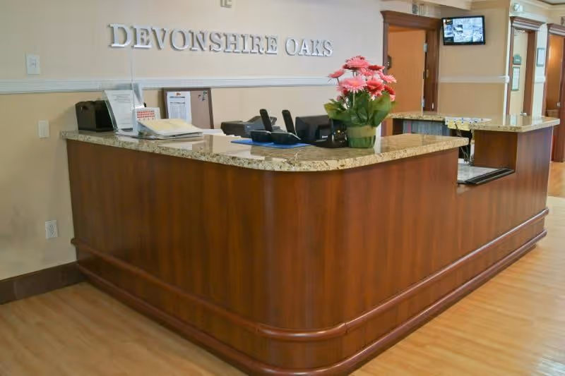 Reception desk at Devonshire Oaks nursing center with a wooden counter, granite countertop, pink flowers, and the facility name on the wall.