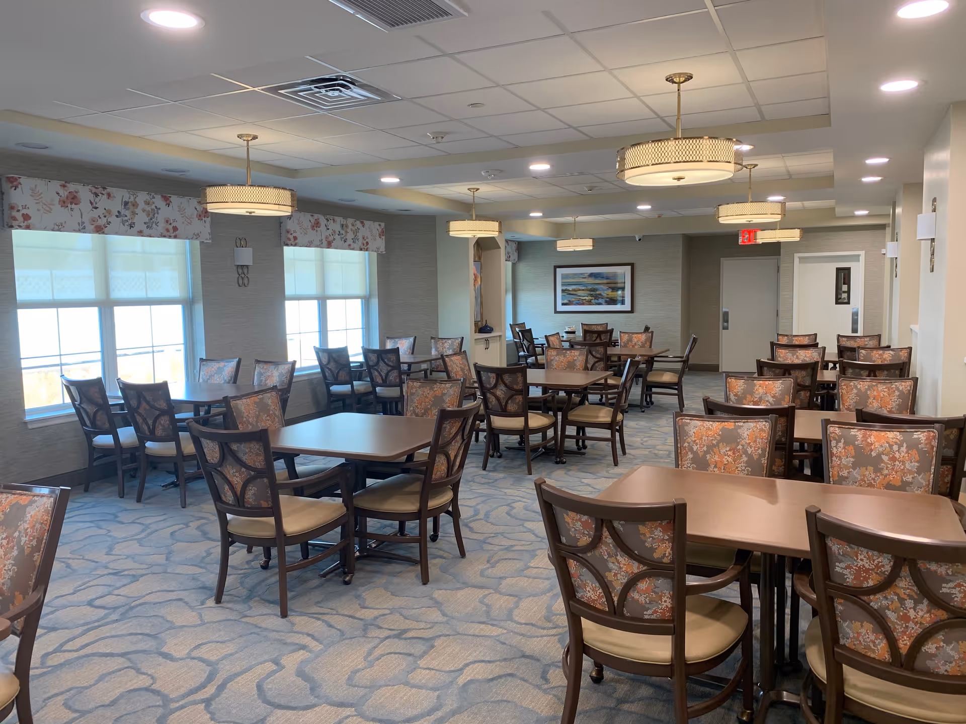 A spacious dining room with multiple wooden tables and chairs featuring floral upholstery. The room has large windows with floral valances, soft carpet with a subtle pattern, and modern ceiling lights. A framed landscape painting is visible on the far wall.