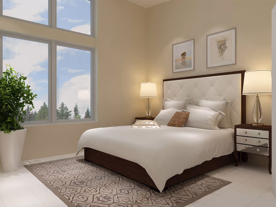 A bright bedroom with a large bed featuring a white tufted headboard, white bedding, and multiple pillows. There are two bedside tables with matching lamps on each side of the bed. Two framed pictures hang on the wall above the headboard. A large window shows a view of trees and a partly cloudy sky. A patterned area rug is placed under the bed, and a tall green plant in a white pot is positioned near the window.