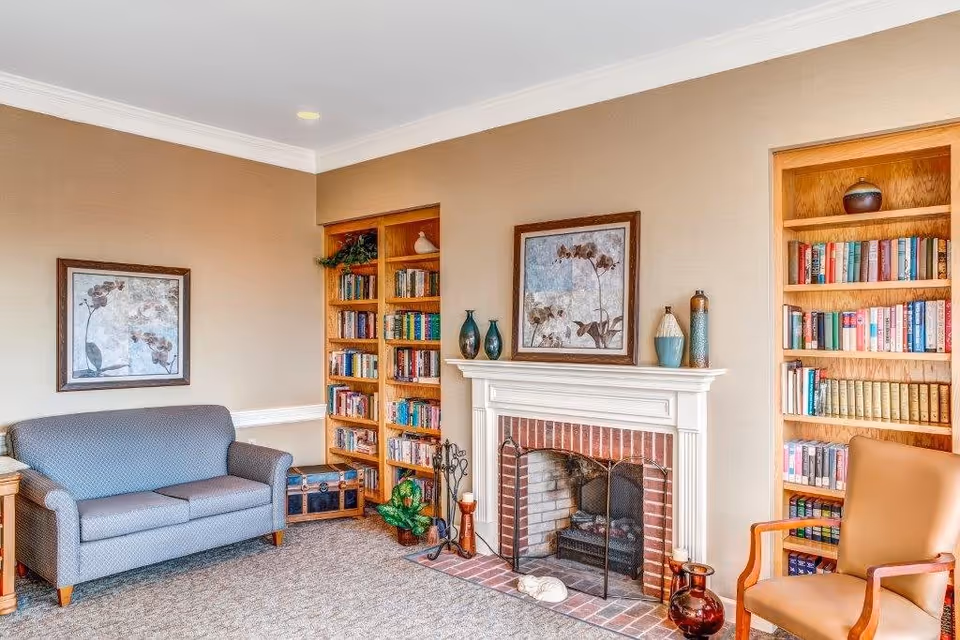A cozy living room area with a gray upholstered loveseat on the left, a brick fireplace with a white mantel in the center, and two wooden bookshelves filled with books on either side of the fireplace. There are decorative vases on the mantel and a framed floral artwork above it. A tan armchair is positioned on the right side of the image. The room has beige walls and carpeted flooring.