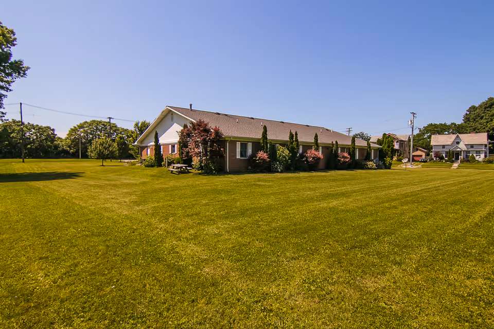 A single-story brick building with a sloped roof surrounded by a large, well-maintained grassy lawn. There are several small trees and shrubs planted along the side of the building. The sky is clear and blue, and there are a few other houses visible in the background.