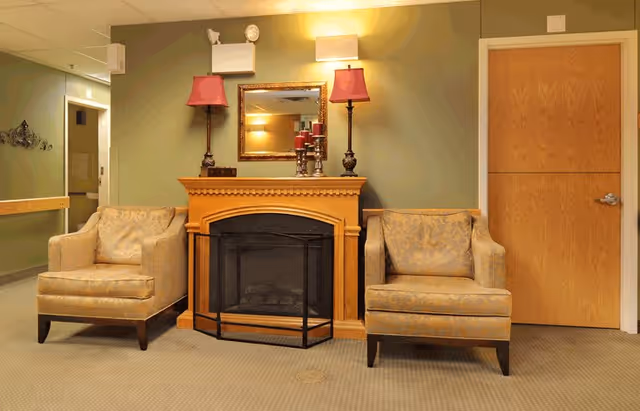 A cozy interior seating area featuring two upholstered armchairs placed on either side of a wooden fireplace with a black screen. Above the fireplace is a rectangular mirror flanked by two red lamps on ornate stands and a set of decorative candles. The walls are painted green, and there is a wooden door to the right and a hallway to the left.
