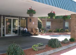 Entrance area of Pine Ridge Health & Rehab facility with a covered drop-off zone. Two people are seated near the entrance, one in a wheelchair. There are hanging flower pots under the canopy and some landscaping with bushes and plants near the building.