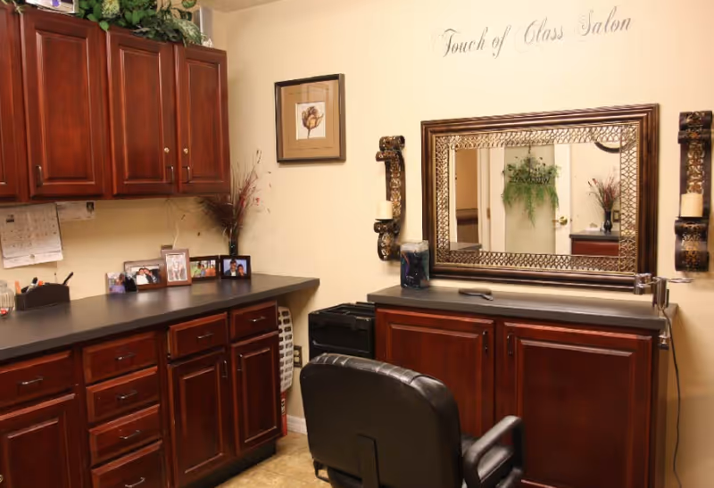 Interior view of a salon room with wooden cabinets, a large decorative mirror on the wall, a black salon chair, and various framed photos and decorative items on the countertops. The wall above the mirror has the text 'Touch of Class Salon'.
