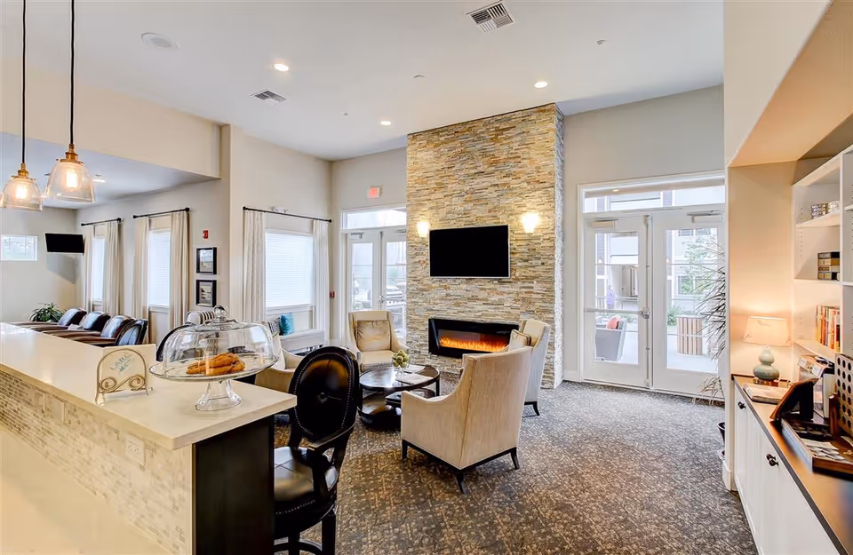 A bright and cozy living room area in a senior living facility featuring a stone fireplace with a mounted flat-screen TV above it. There are several comfortable chairs arranged around a small round table in front of the fireplace. To the left, there is a counter with a glass dome containing cookies and a sign labeled 'LARC'. The room has large windows with curtains and glass doors leading outside. Shelves with books and a lamp are visible on the right side.