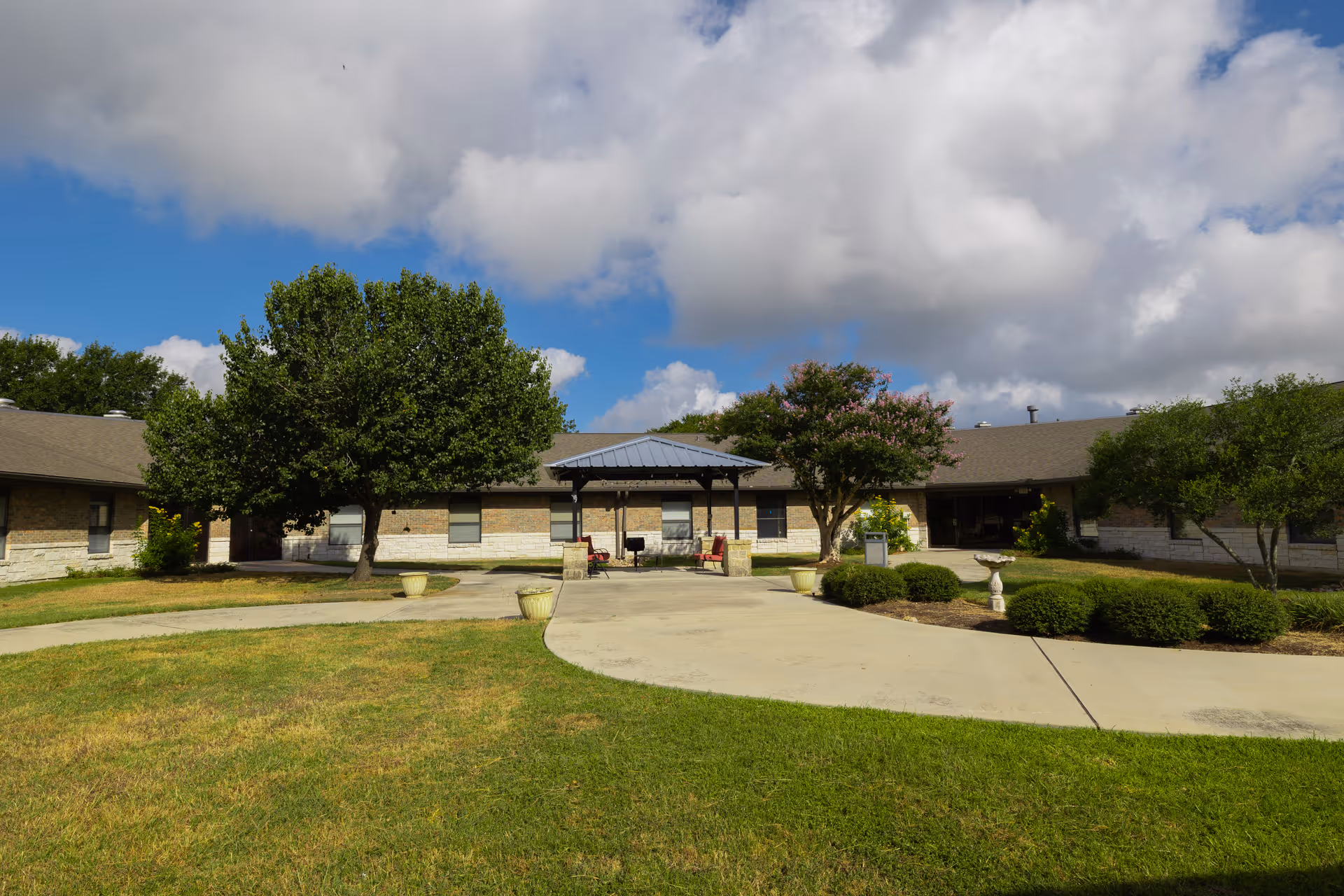 Exterior front entrance of a single-story nursing facility with a covered entry, trees, and a circular driveway under a partly cloudy sky.