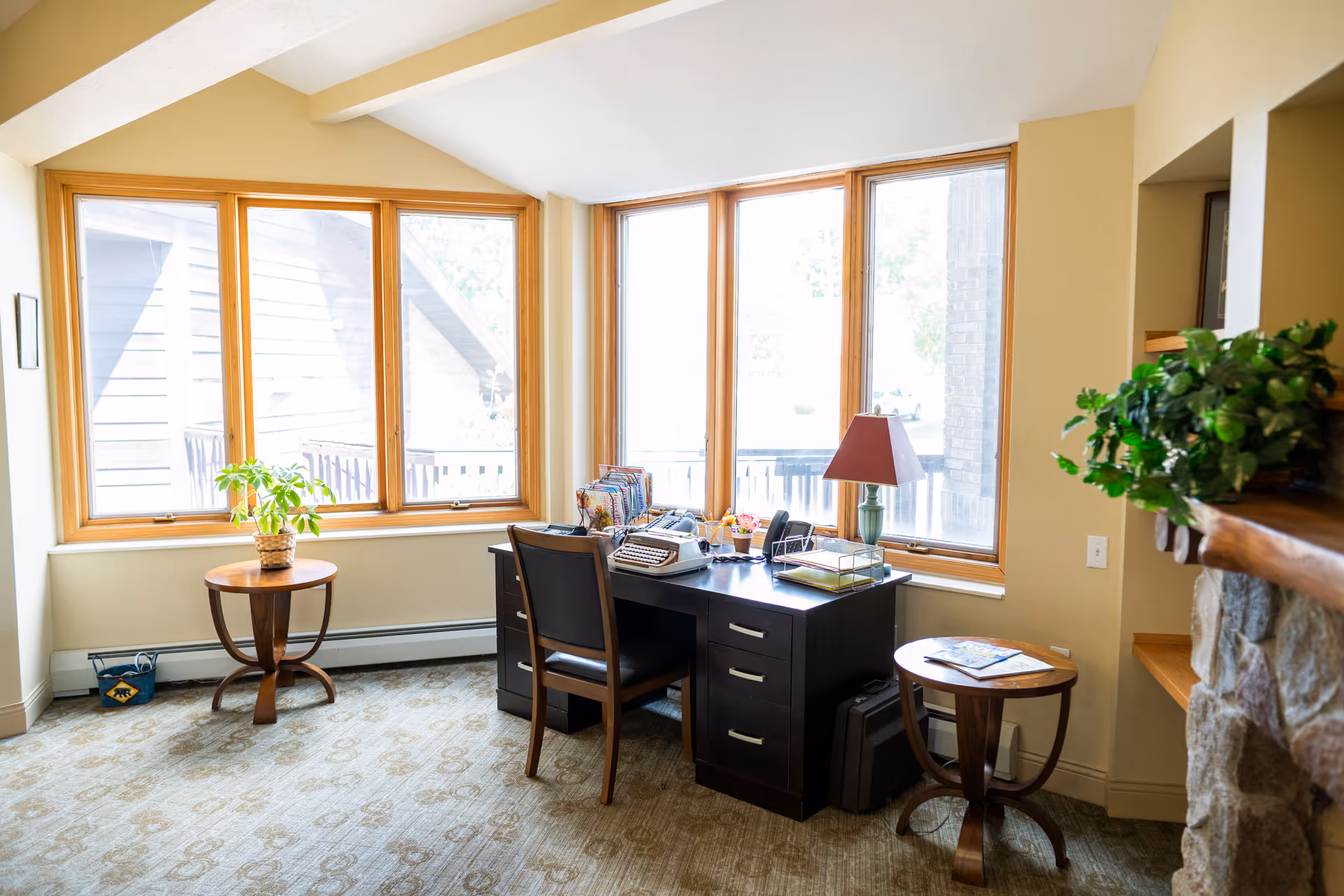 A bright room with large windows letting in natural light, featuring a dark wooden desk with a chair, a typewriter, a lamp, and office supplies. There are two small round wooden tables on either side of the desk, one with a potted plant and the other with magazines. The room has beige walls, a patterned carpet, and a stone fireplace partially visible on the right.