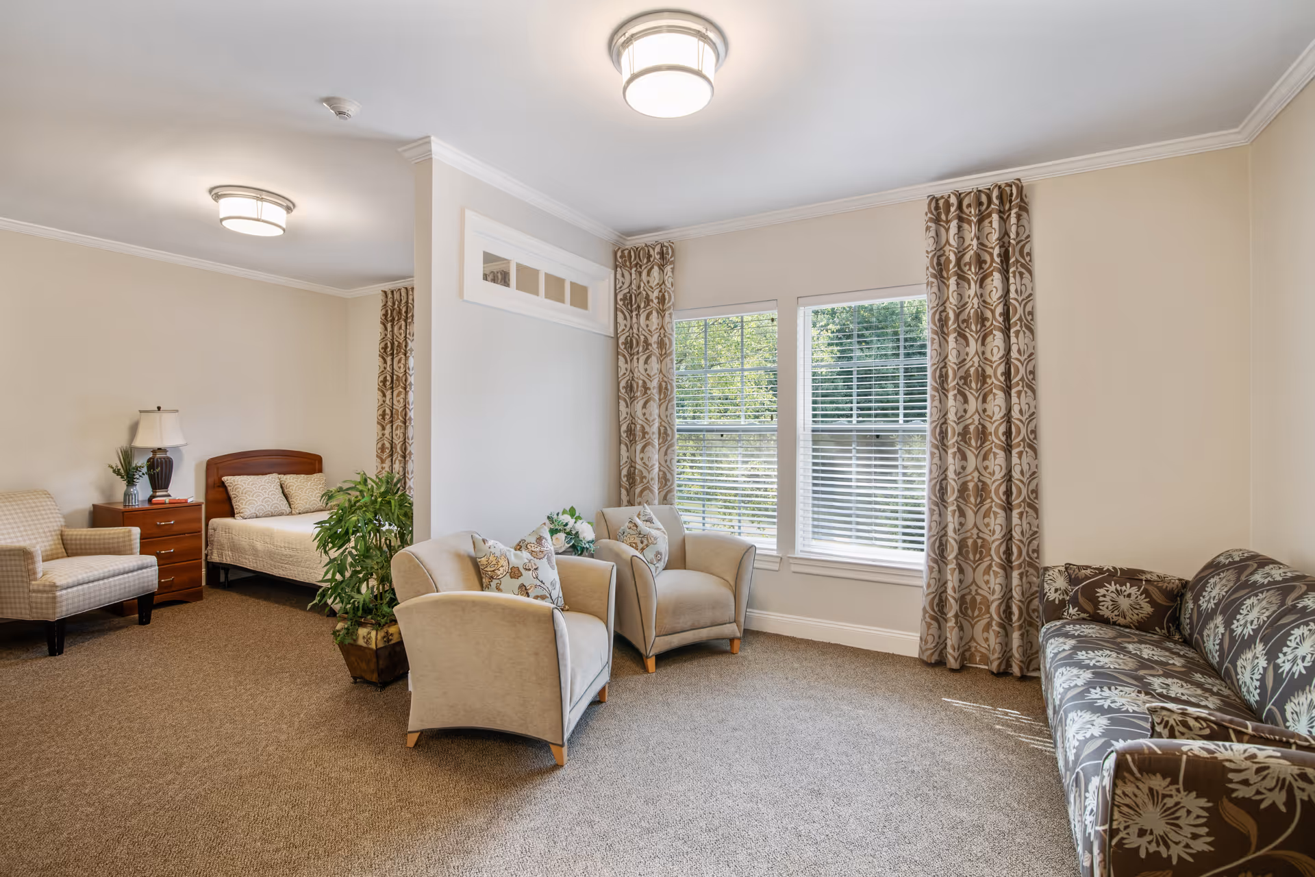 A bright and cozy room in a senior living facility featuring a sitting area with two beige armchairs with floral cushions, a patterned sofa, and large windows with patterned curtains letting in natural light. In the background, there is a sleeping area with a bed, a wooden nightstand with a lamp, and an armchair. The room has beige walls and carpeted flooring.