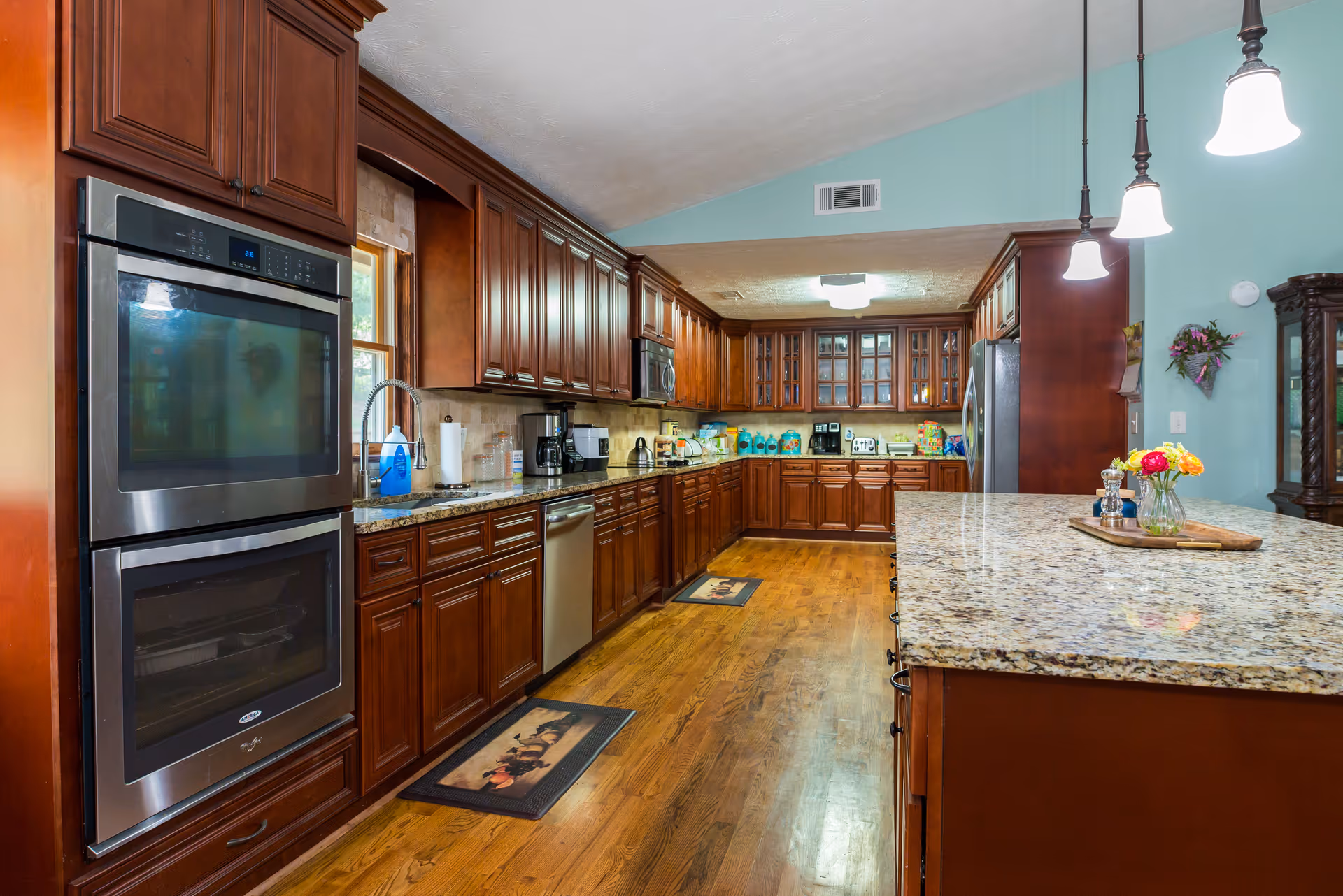 A spacious kitchen with wooden cabinets and hardwood floors. The kitchen features a double oven, a stainless steel dishwasher, a microwave, and a refrigerator. There is a large granite countertop island with pendant lights hanging above it. Various kitchen appliances and containers are placed on the countertops, and a small vase with colorful flowers is on the island. The walls are painted light blue.