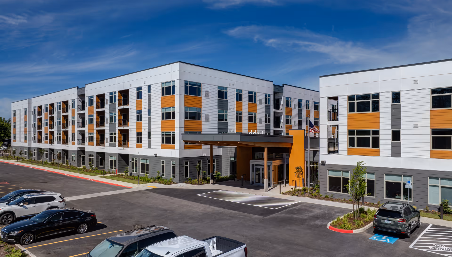 Front exterior of a modern four-story senior living building with a covered entrance, balconies, and a parking lot.
