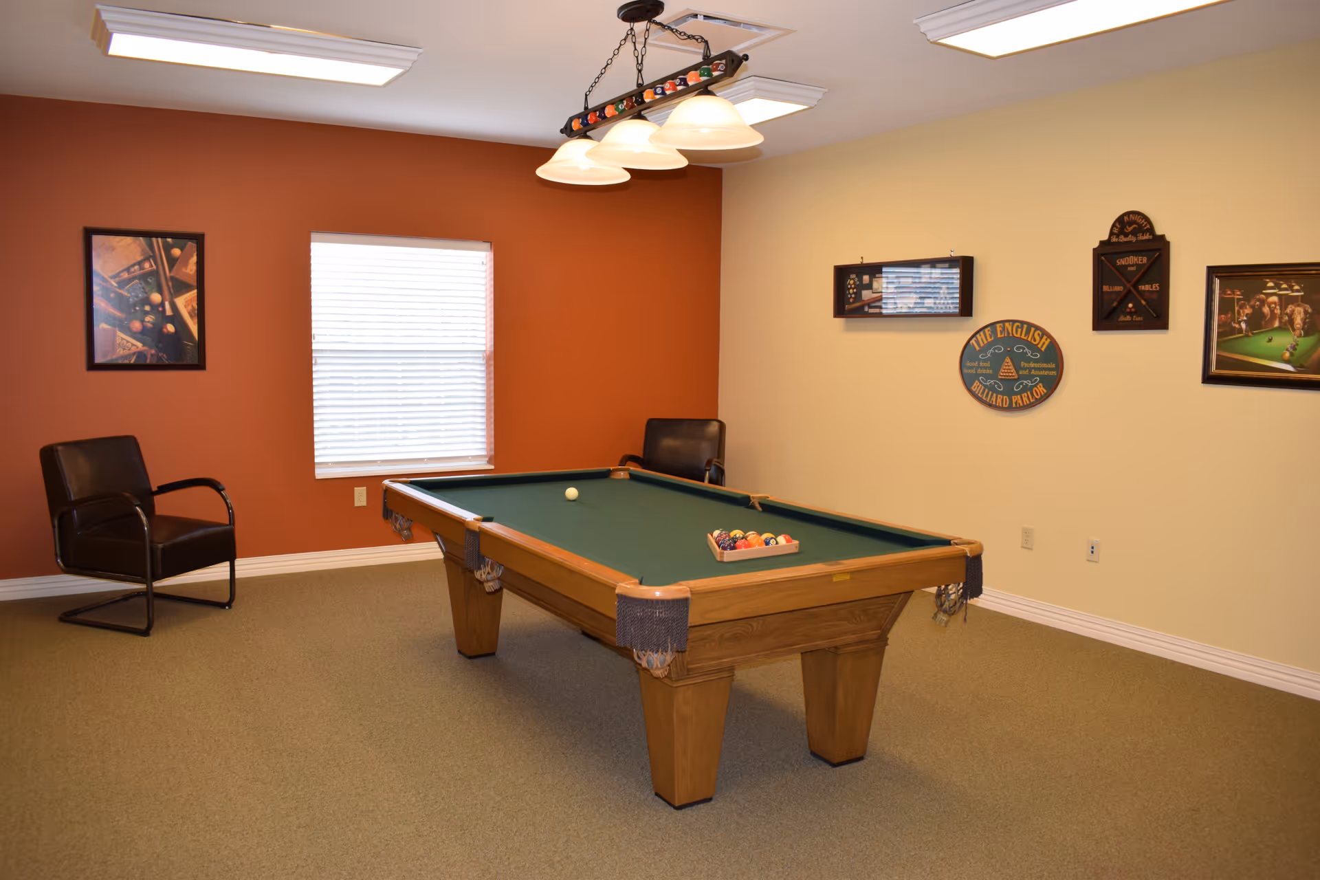A game room with a pool table in the center, two black chairs against the walls, a window with blinds, and several billiards-themed pictures and signs on the walls. The walls are painted in two colors: one side is a warm brown and the other is a light cream color. A hanging light fixture with four lamps is above the pool table.