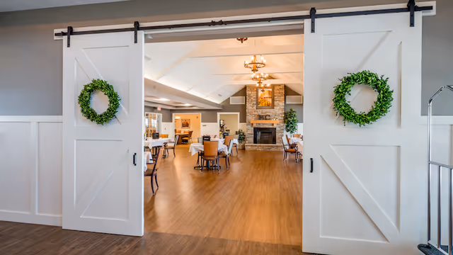 View through large white sliding barn doors decorated with green wreaths into a spacious dining room with wooden floors, tables and chairs set for meals, and a stone fireplace at the far end under a vaulted ceiling with chandeliers.