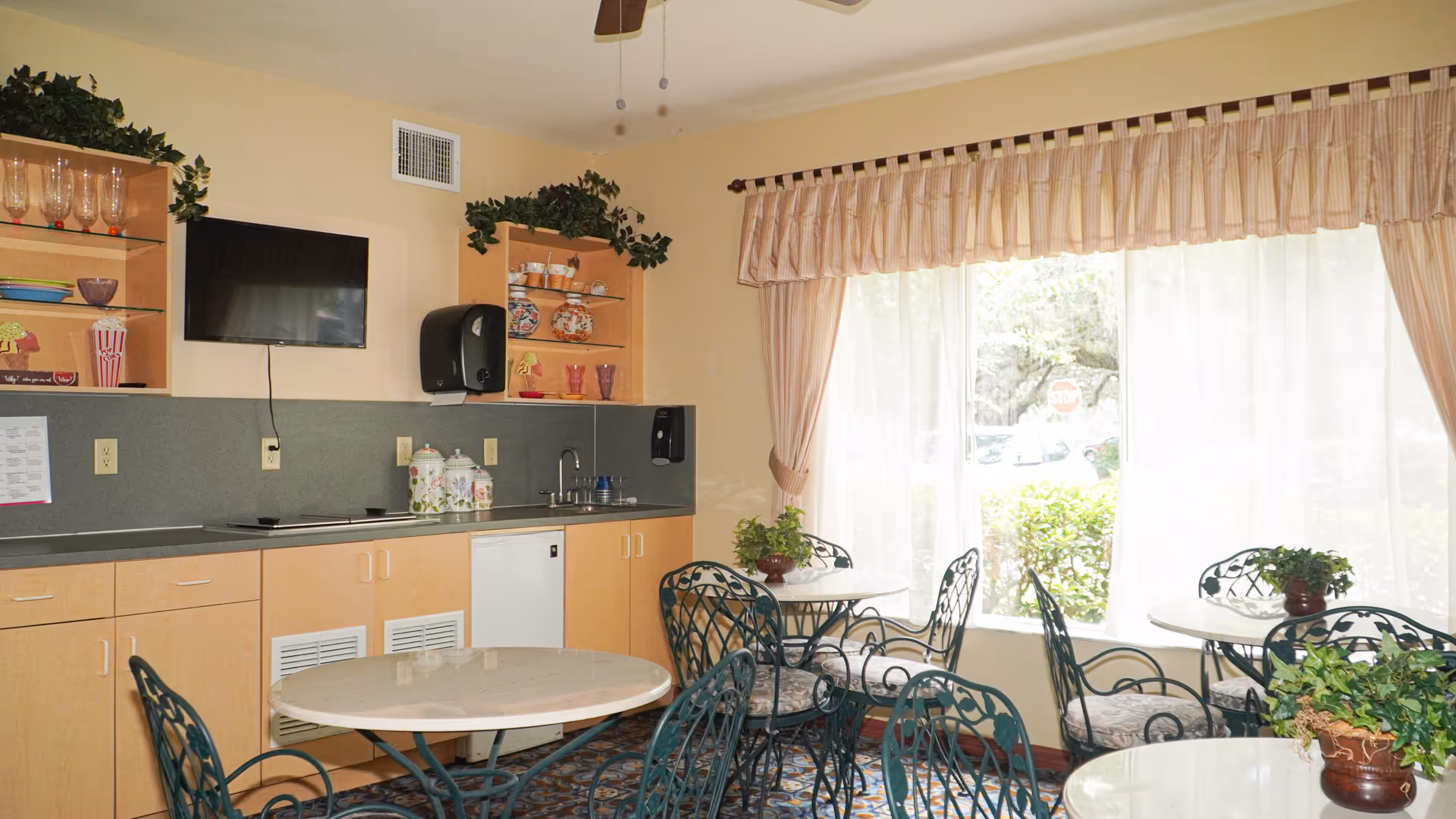 Bright communal dining area with round tables and wrought-iron chairs beside a kitchenette and large curtained window.