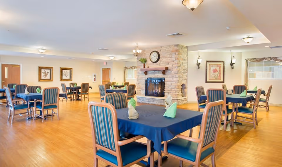 Spacious dining room with multiple tables and striped chairs, navy tablecloths and folded napkins, and a central stone fireplace with a clock.