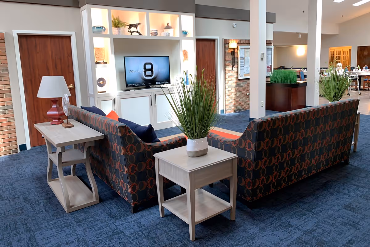 A cozy common area in a senior living facility with two patterned sofas arranged around a small wooden table with a potted plant. There is a TV mounted on a white built-in shelving unit displaying a screen with the number 8. The room has blue carpet, wooden doors, and a mix of brick and beige walls. Additional seating and tables are visible in the background.