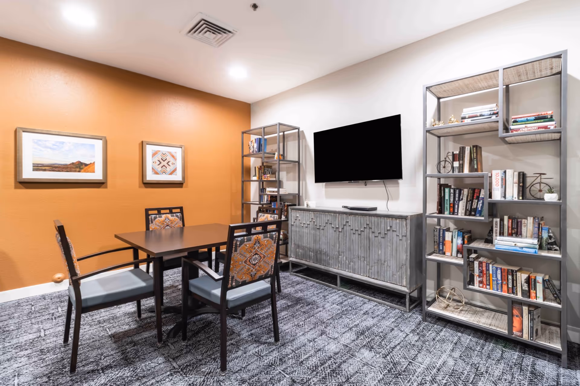 A cozy interior room with a square table surrounded by four chairs with patterned cushions. The room features a burnt orange accent wall with two framed pictures, a mounted flat-screen TV on a white wall, and two metal bookshelves filled with books and decorative items. The floor is carpeted with a patterned gray carpet.