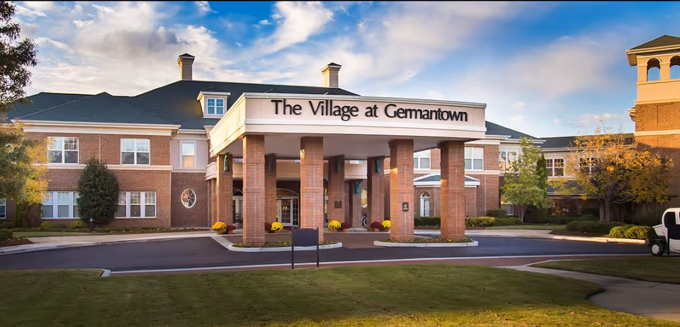 Front exterior view of The Village at Germantown building with a covered entrance supported by brick columns, surrounded by trees and a well-maintained lawn under a partly cloudy sky.
