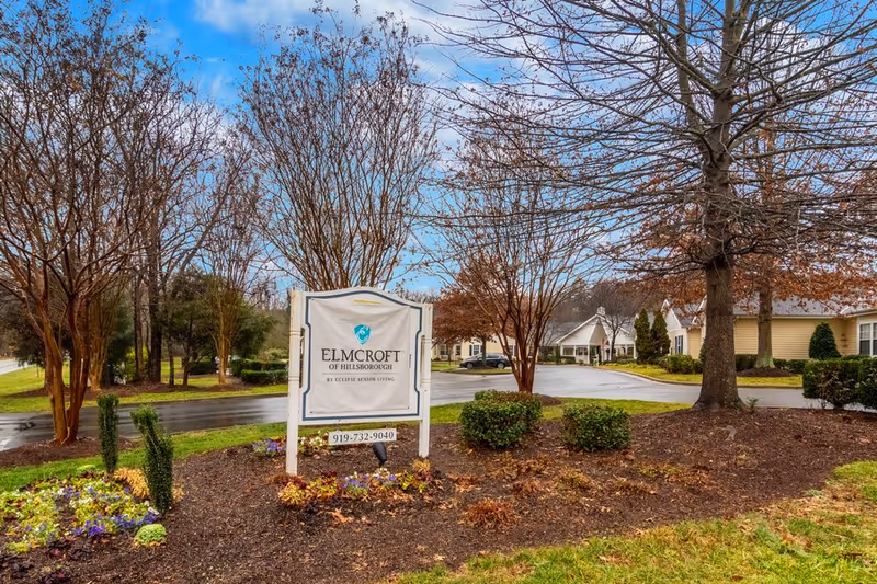 Entrance sign reading "Elmcroft of Hillsborough" surrounded by landscaping with facility buildings and trees in the background on a cloudy day.