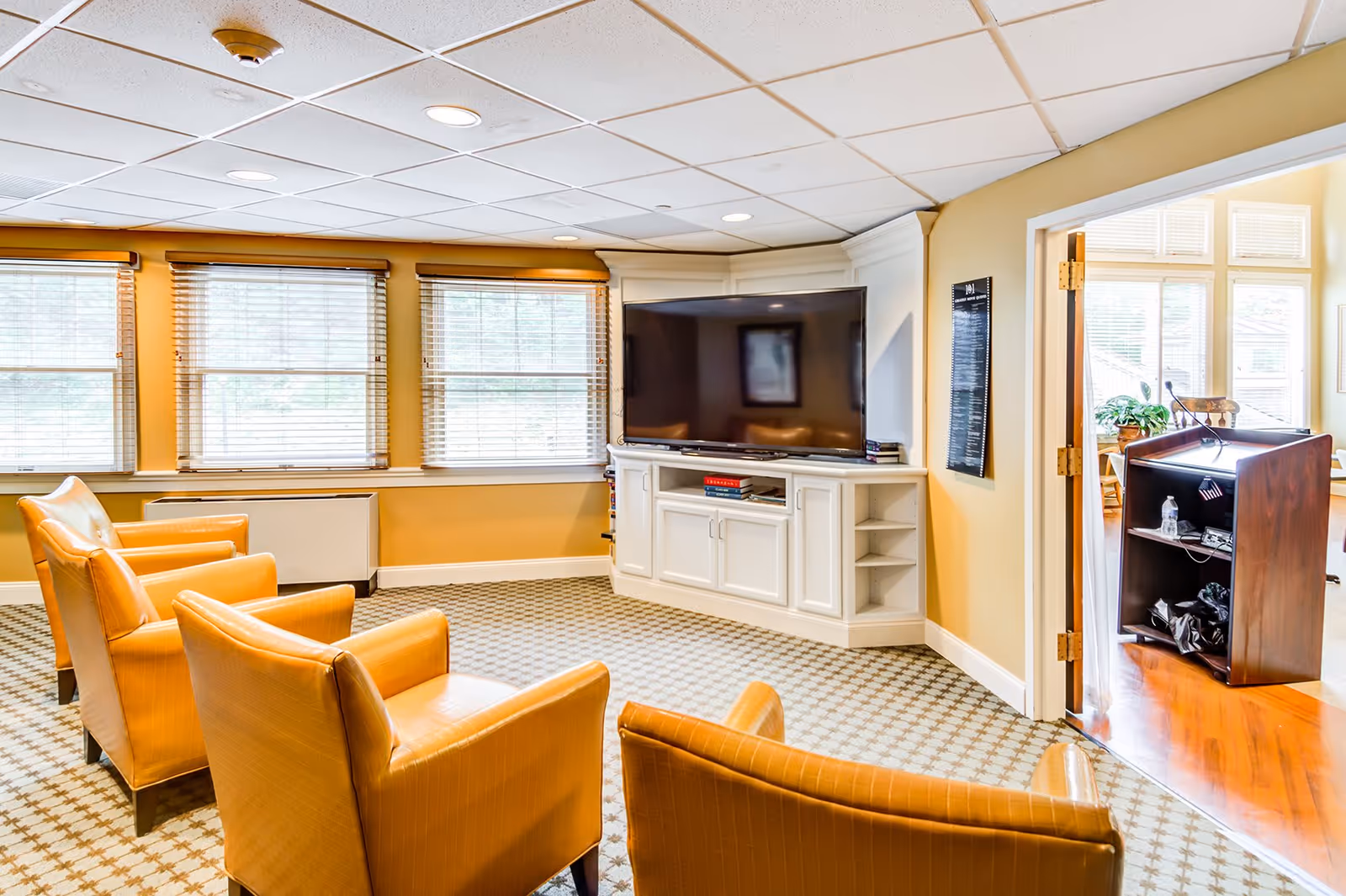 A cozy living room area with four tan leather armchairs arranged in front of a large flat-screen TV mounted on a white built-in cabinet. The room has beige walls, a patterned carpet, and three large windows with blinds letting in natural light. An open doorway leads to another room with wooden flooring and a podium.