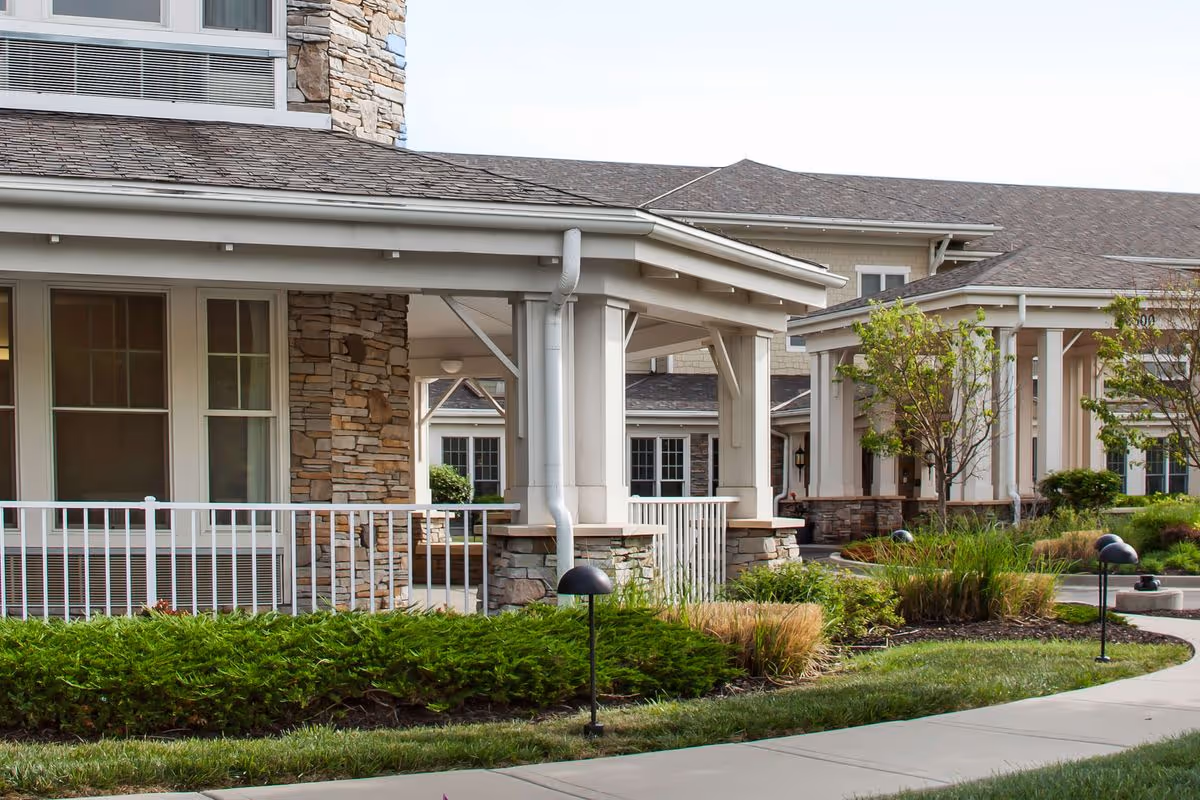 Covered entrance and porch of a senior living building with stone accents, landscaping, and walkways.