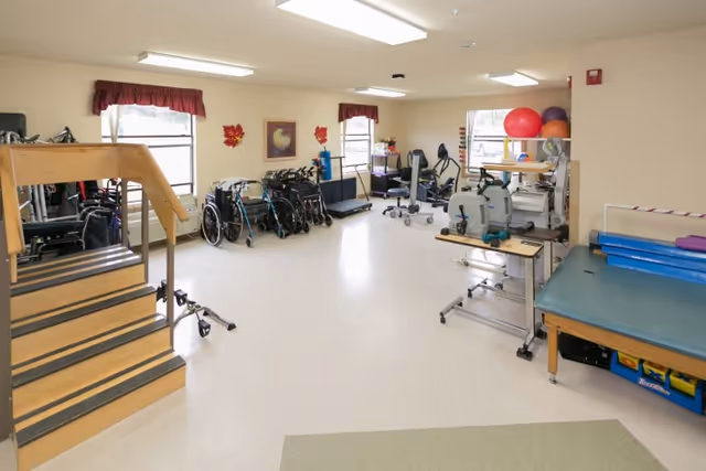 A spacious rehabilitation room with exercise equipment, wheelchairs, walkers, parallel bars, therapy balls, and a small staircase for physical therapy exercises. The room has beige walls, two windows with red valances, and bright overhead lighting.