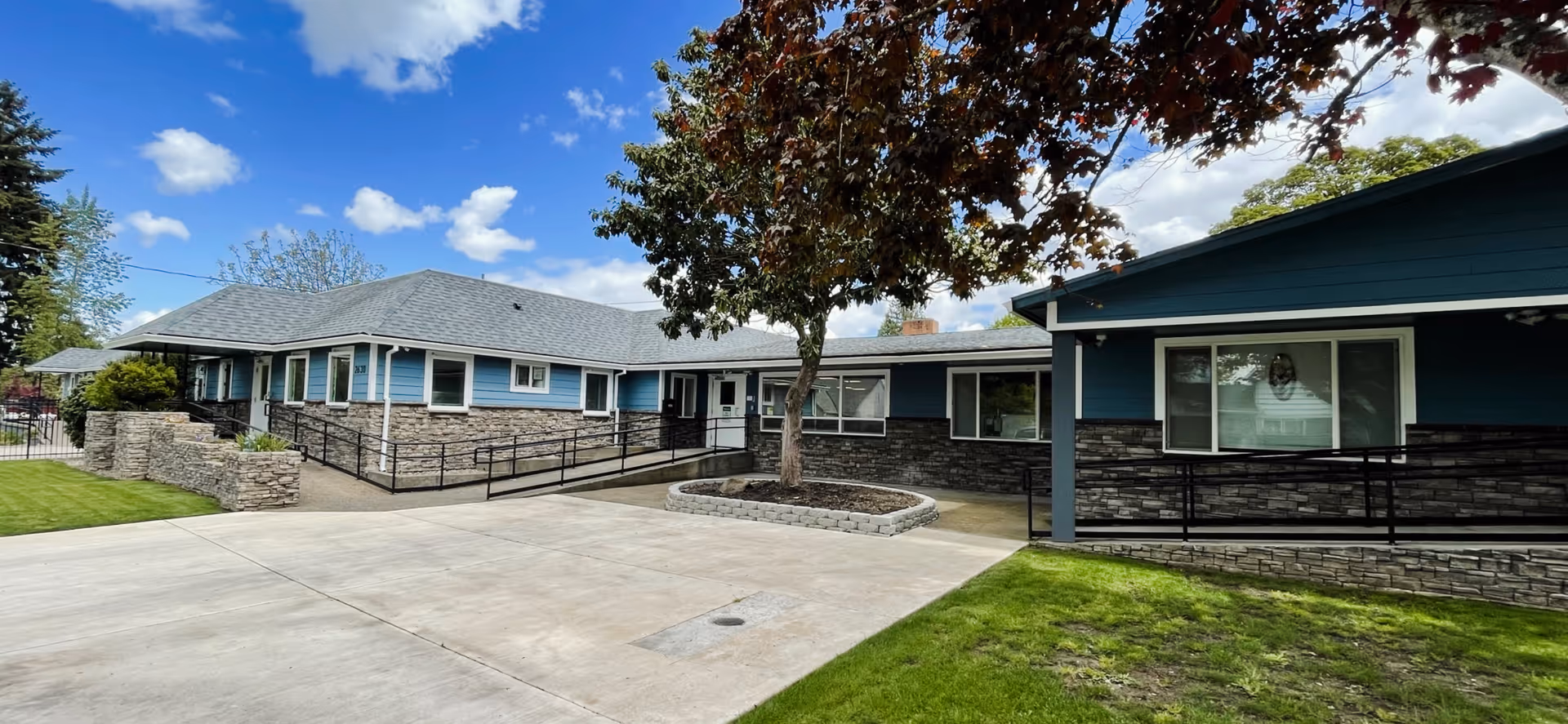 Exterior view of a single-story building with blue siding and stone accents, featuring a concrete pathway, a tree in the center, and a well-maintained lawn under a partly cloudy sky.