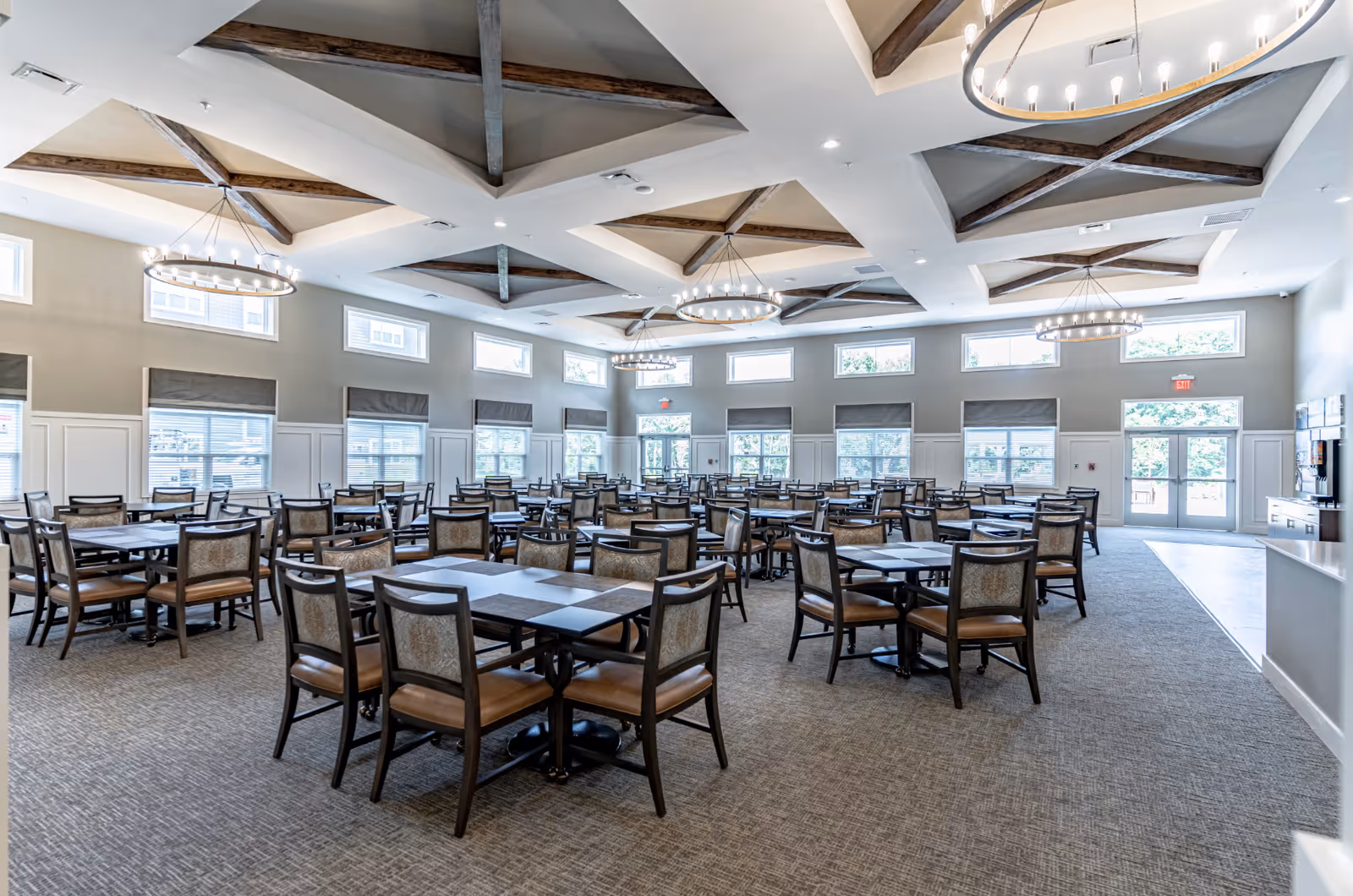 A spacious dining room with multiple square tables and cushioned chairs arranged neatly. The room features large windows with gray shades, a high ceiling with wooden beams, and modern circular chandeliers providing warm lighting. The floor is carpeted, and there is a door leading outside at the far end.