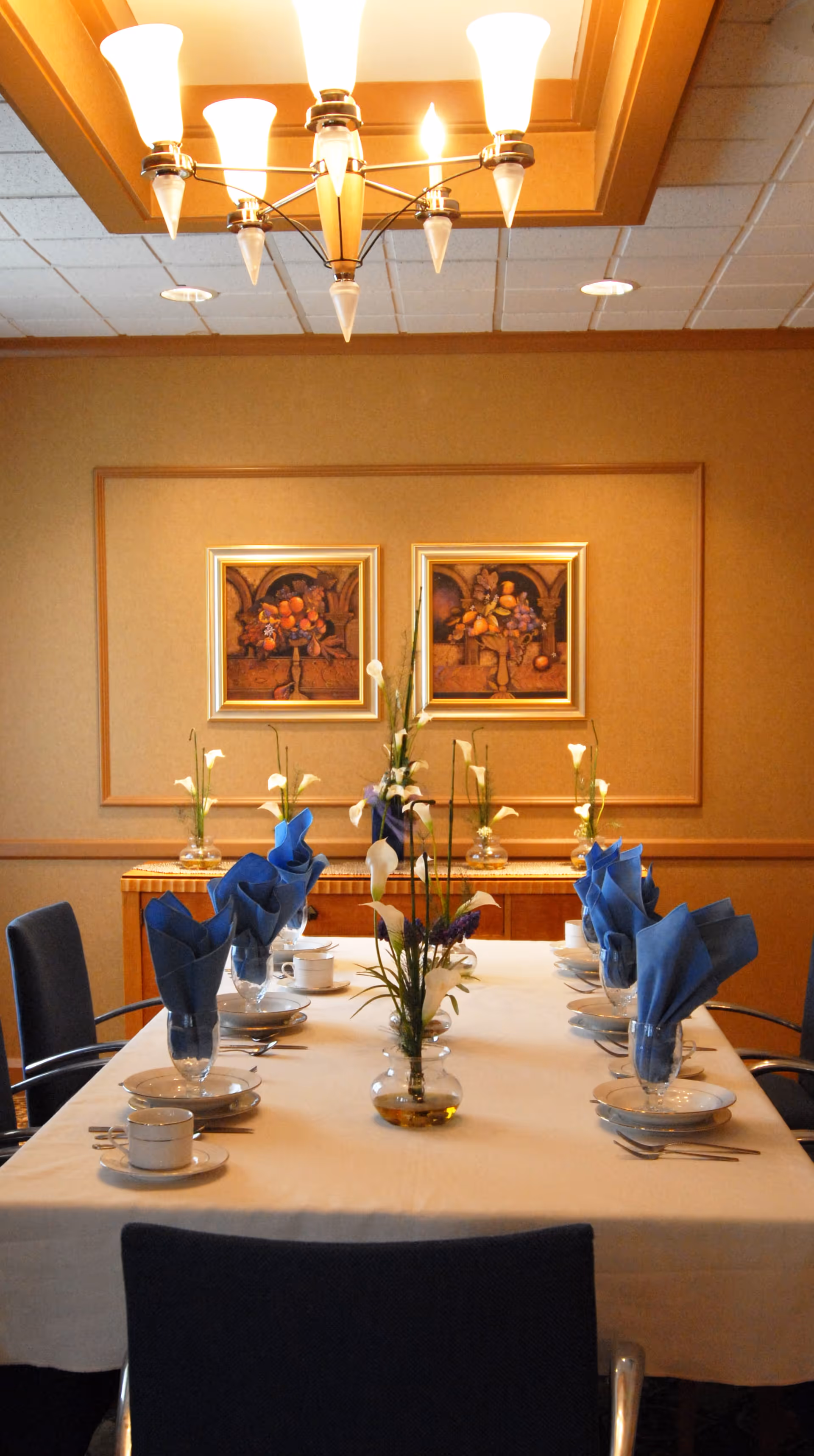 A formal dining room with a rectangular table set for six people. The table is covered with a white tablecloth and has blue folded napkins in glasses, white plates, cups, and silverware. There are flower arrangements in small vases on the table. The room has beige walls with two framed paintings of fruit and a wooden sideboard against the back wall. A chandelier with multiple lights hangs from the ceiling.
