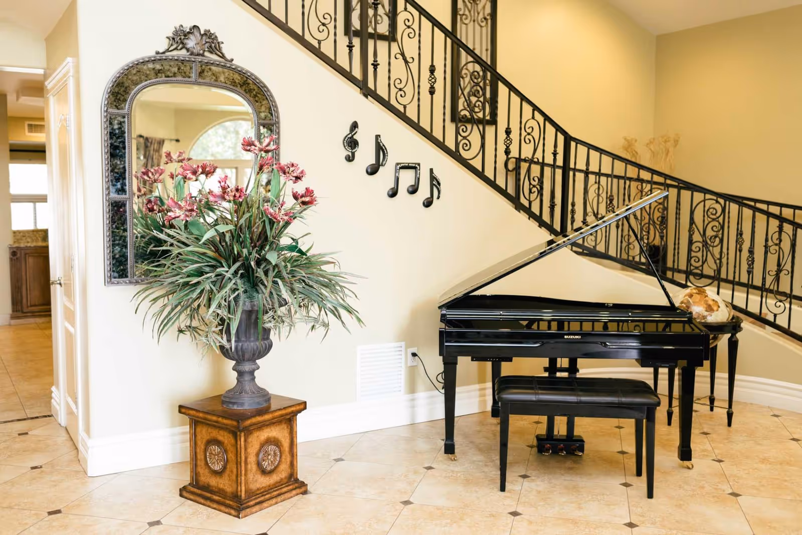 Interior view of a room featuring a black grand piano with a matching bench, a decorative plant in a vase on a wooden pedestal, a large ornate mirror on the wall, and a staircase with wrought iron railing adorned with musical note decorations.