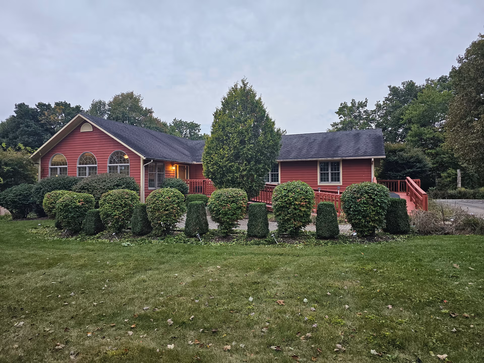 A single-story red building with a dark roof surrounded by neatly trimmed bushes and a green lawn. The building has three arched windows on the left side and a ramp leading to the entrance. Trees and greenery are visible in the background under a cloudy sky.