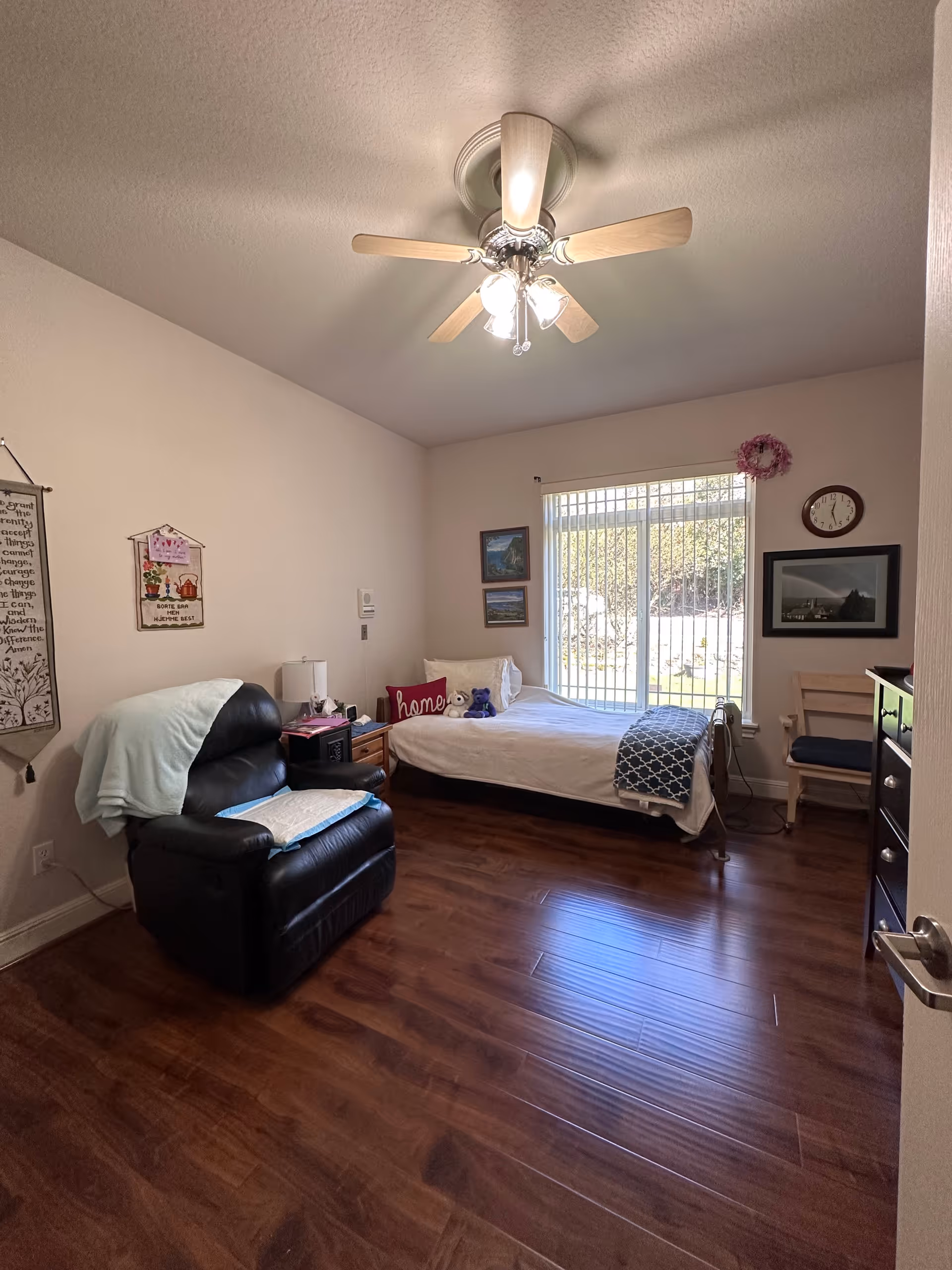 A tidy bedroom with a single bed, a black recliner, a ceiling fan, wooden floors and a large window with vertical blinds.