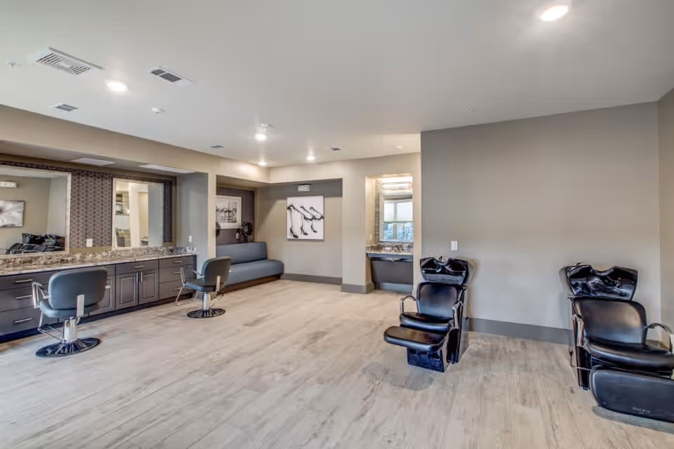 Interior view of a salon area in a senior living facility with two black hair washing stations on the right, two styling chairs in front of a long counter with mirrors on the left, and a gray bench seating area in the back corner. The room has light wood flooring and neutral-colored walls with recessed lighting.