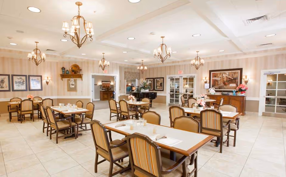 A spacious dining room with multiple tables and chairs arranged neatly. The room features beige tiled floors, striped wallpaper, and several chandeliers hanging from the ceiling. There are framed pictures on the walls and floral centerpieces on the tables. Double doors and an upright piano are visible in the background.