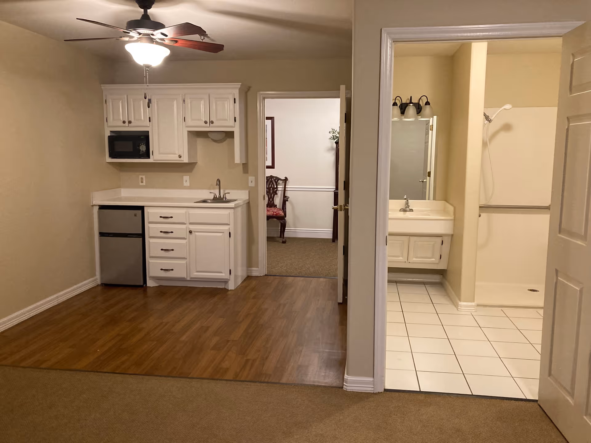 Studio-style interior showing a small kitchenette with microwave and mini-fridge, ceiling fan, and an adjacent bathroom with sink and shower.