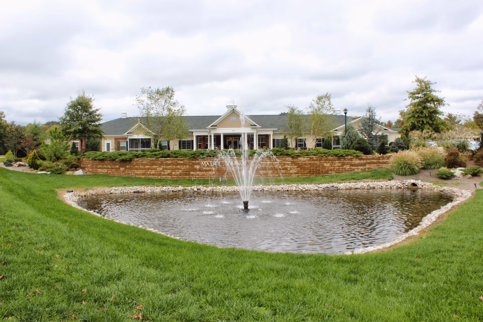 Exterior view of a senior living facility with a pond and a water fountain in the foreground, green grass surrounding the pond, and a building with a covered entrance and columns in the background under a cloudy sky.