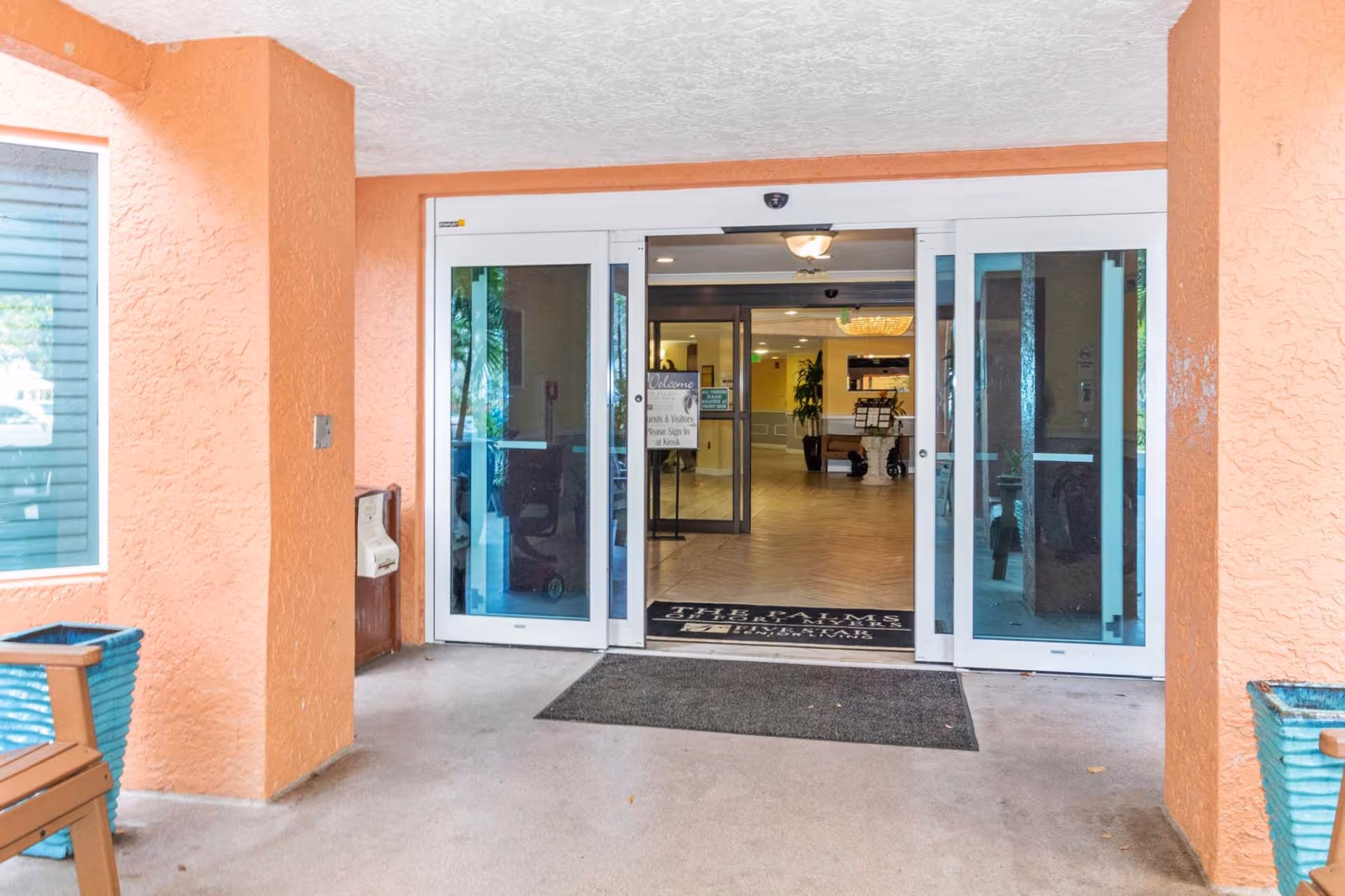 Entrance to The Palms of Fort Myers facility showing automatic sliding glass doors, peach-colored textured walls, a welcome mat with the facility name, and a glimpse of the interior lobby area with plants and seating.