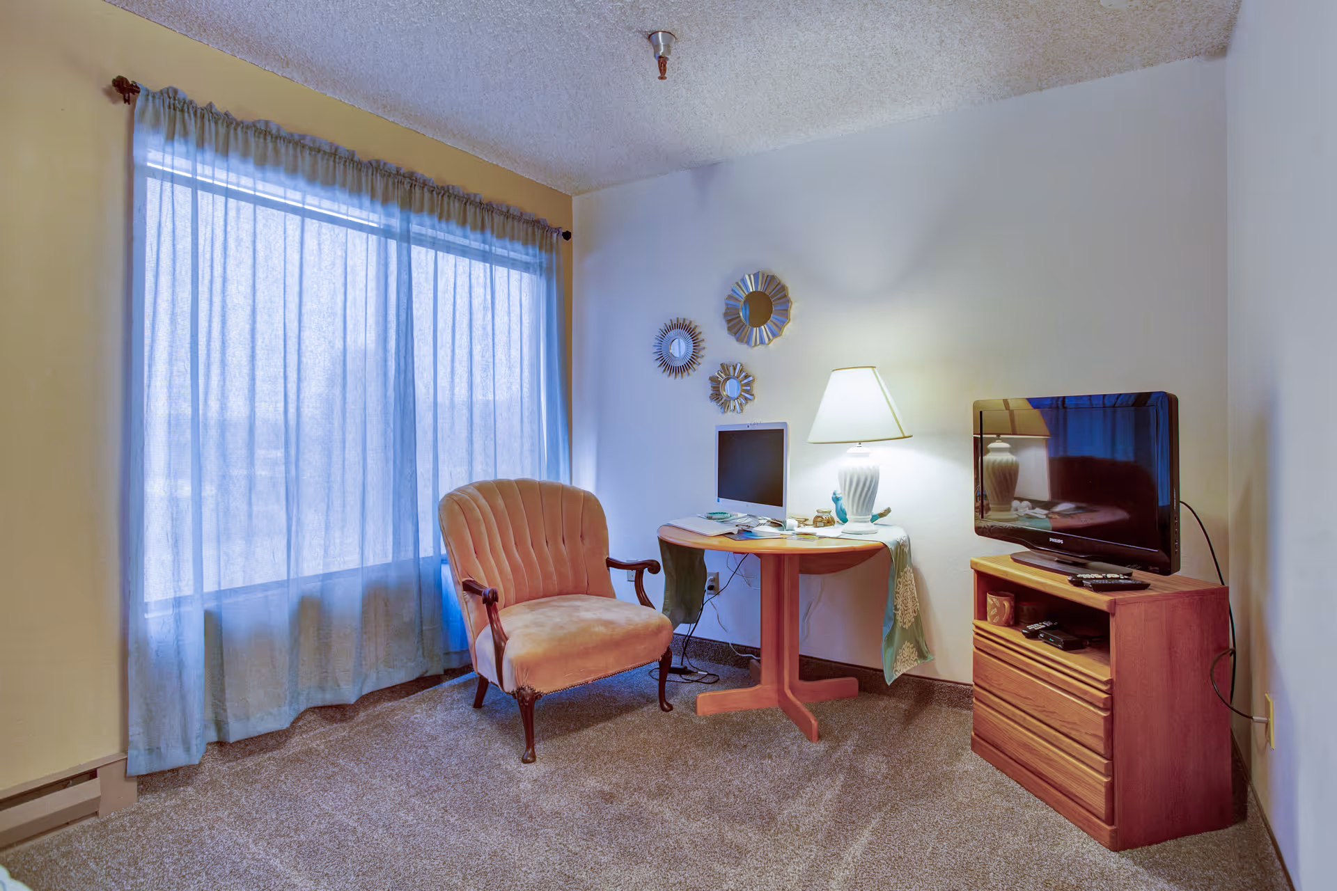 A cozy living room area with a large window covered by sheer curtains, a pink upholstered armchair, a round wooden table with a lamp and computer monitor on it, and a wooden TV stand with a flat-screen television and remote controls.
