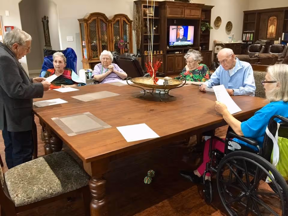 A group of elderly residents sit around a large wooden table in a common room, reading papers with a TV and cabinetry in the background.