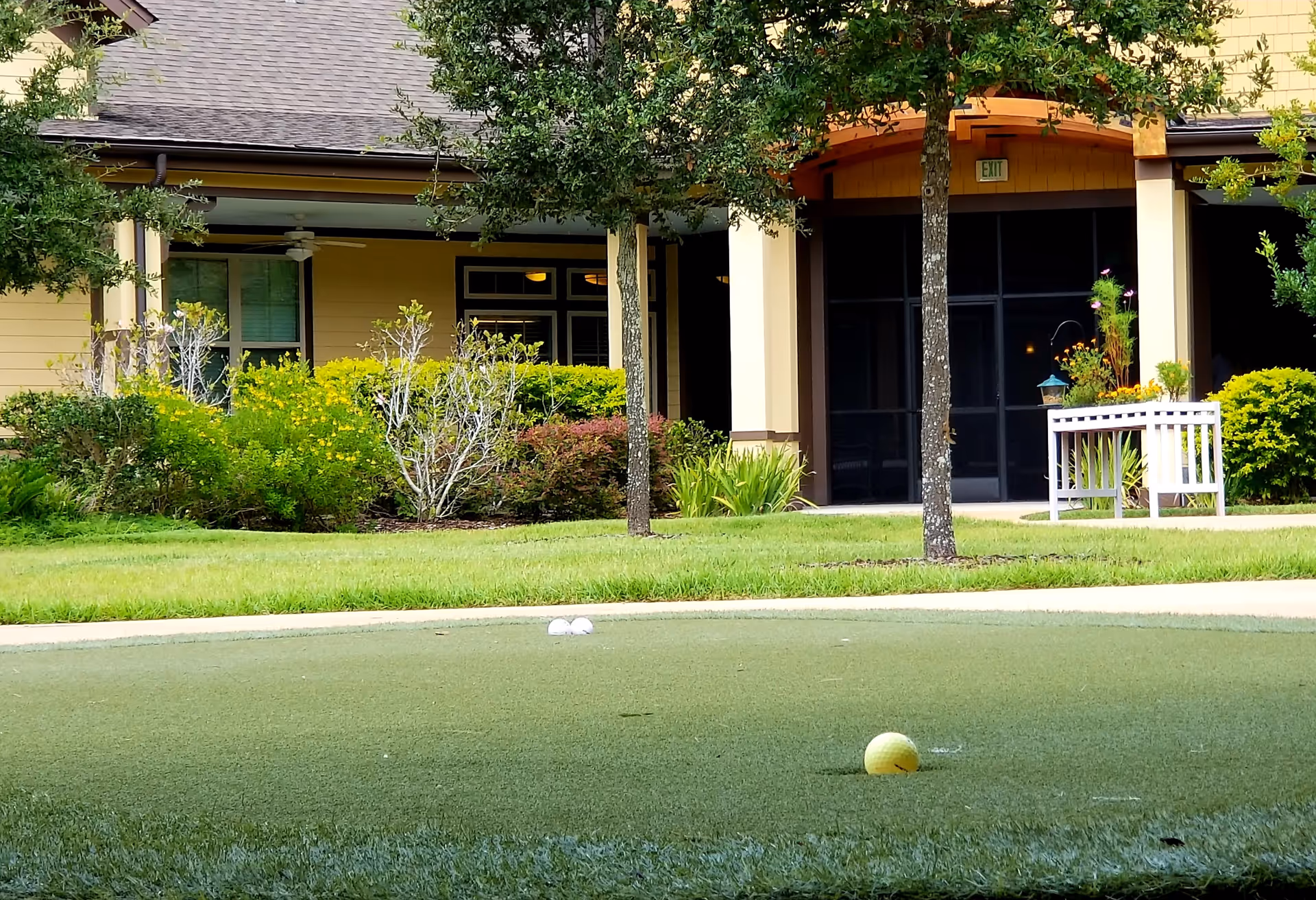 Outdoor putting green with golf balls in the foreground, surrounded by grass and trees, with a building entrance and landscaping visible in the background.