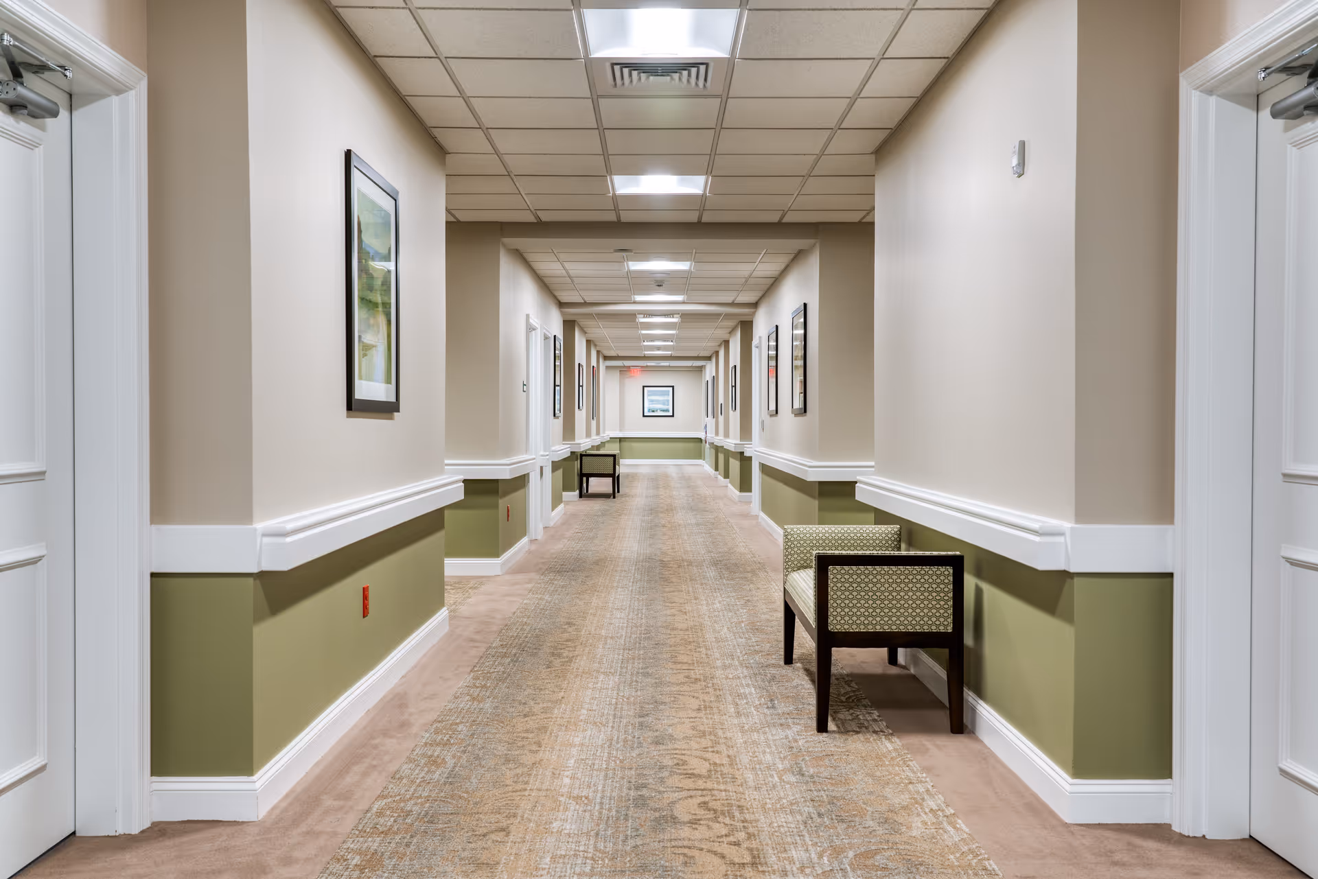 A long, well-lit hallway in a senior living facility with beige and green walls, carpeted floor, framed artwork on the walls, and cushioned benches along the sides.