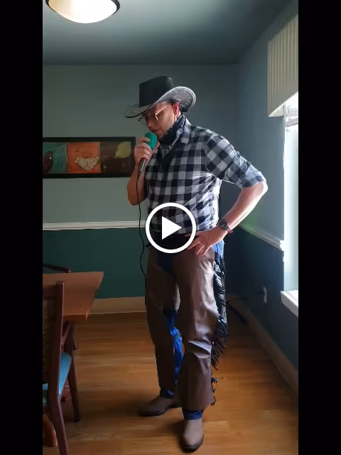 A person dressed in a cowboy outfit, including a black cowboy hat, checkered shirt, brown chaps, and boots, is standing indoors holding a microphone near their mouth. The room has wooden flooring, green and white walls, a window with blinds, a ceiling light, and a framed artwork on the wall.
